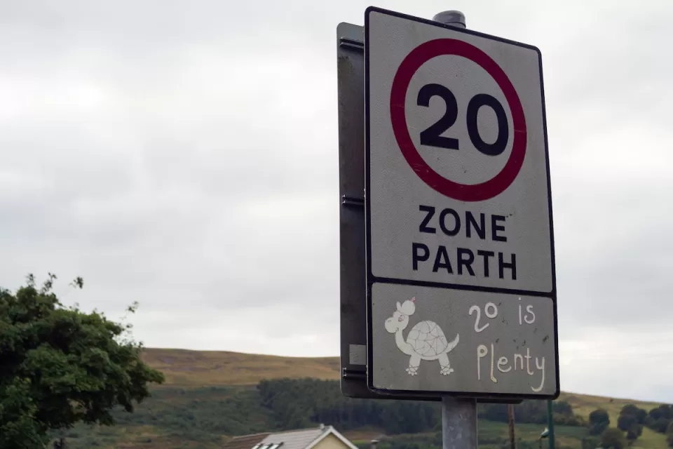 Close-up of white 20 kph speed limit sign in Europe with cartoon turtle and '20 is plenty' sticker underneath.